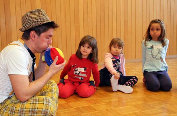 Ein Clown bläst einen bunten Ball vor drei aufmerksamen Mädchen auf.