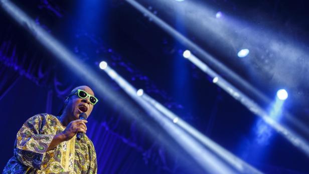 Stevie Wonder singt mit grüner Sonnenbrille auf einer Bühne mit blauem Licht.