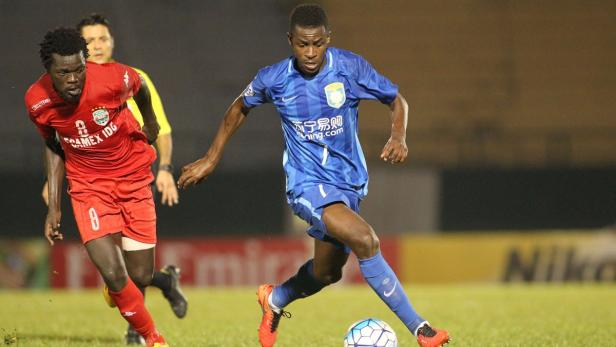 Ein Fußballspieler in einem blauen Trikot dribbelt den Ball, während ein Gegenspieler in Rot versucht, ihn aufzuhalten.