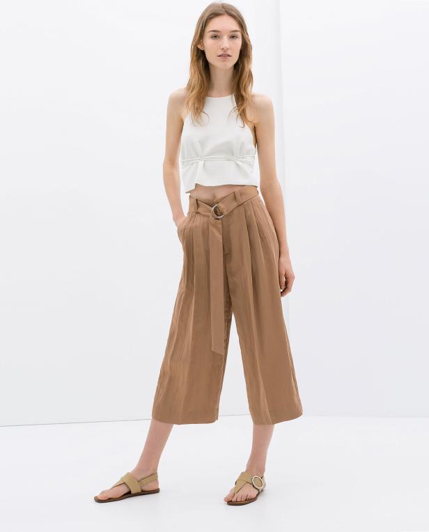 Eine Frau posiert in einem weißen Top und einer braunen Culotte mit Gürtel.