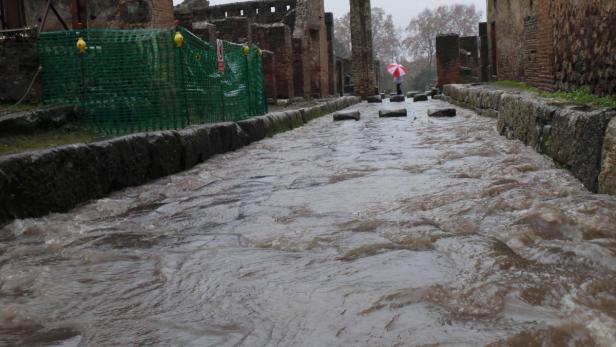 Ein überfluteter Weg in Pompeji mit einer Person mit Regenschirm im Hintergrund.