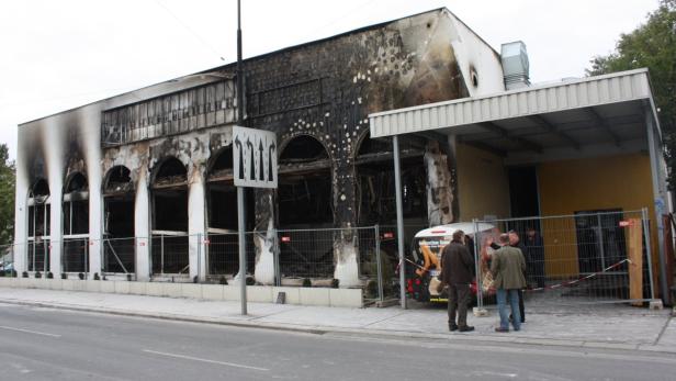 Ein ausgebranntes Gebäude mit verkohlter Fassade und mehreren Personen davor.