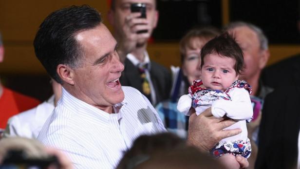 Mitt Romney hält ein Baby im Arm und lächelt.