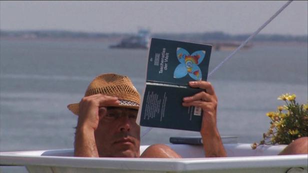 Ein Mann mit Hut liest in einer Badewanne sitzend einen „Taschenatlas der Welt“.