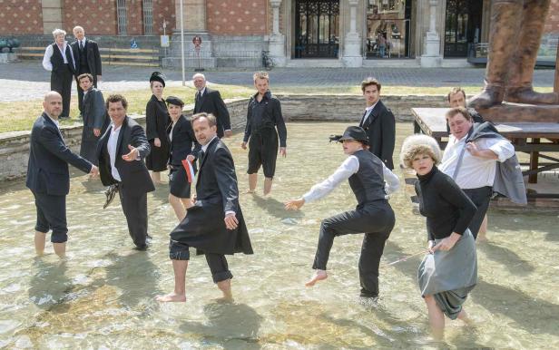 Eine Gruppe von Menschen watet barfuß in einem flachen Wasserbecken vor einem Gebäude.