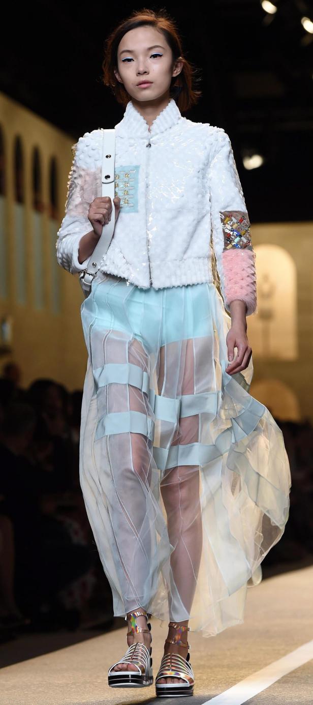 Ein Model präsentiert ein weißes Jackett und einen transparenten Rock auf dem Laufsteg.