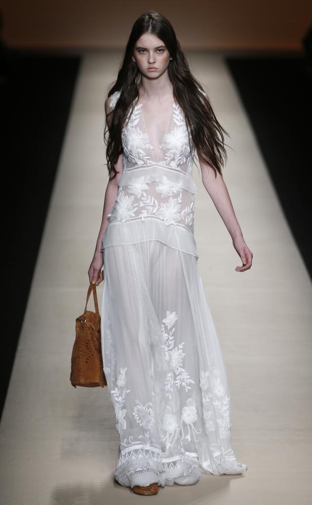 Ein Model präsentiert ein weißes, transparentes Kleid mit Blumenmuster und eine braune Tasche auf dem Laufsteg.