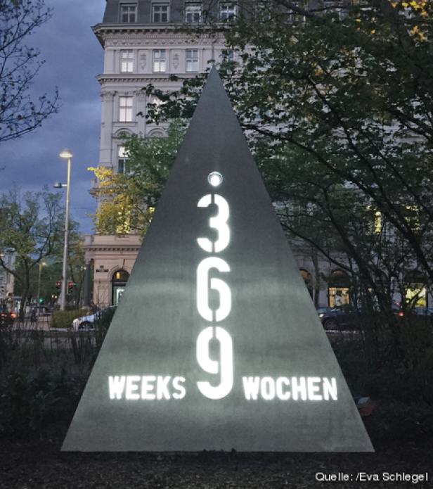Eine dreieckige Skulptur mit der Aufschrift „369 Weeks Wochen“ steht in einem Park.
