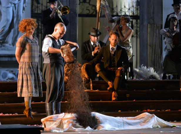 Eine Theaterszene mit Musikern und einem Mann, der Erde auf eine unter einem Tuch liegende Person schüttet.