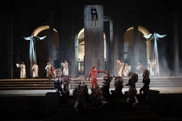 Eine Theaterszene mit Engeln, Musikern und Personen mit Dämonenmasken auf einer Bühne.