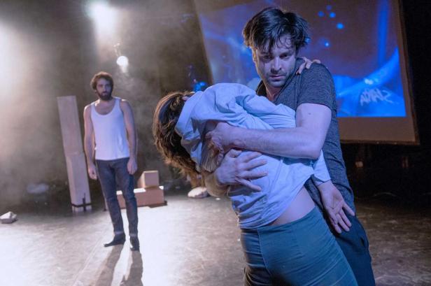 Auf einer Bühne umarmt ein Mann eine andere Person, während ein dritter Mann im Hintergrund steht.