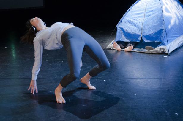 Eine Frau mit Kopfhörern tanzt vor einem blauen Zelt, aus dem Füße ragen.