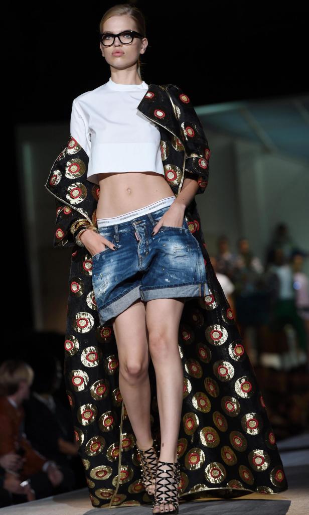 Ein Model mit Brille, bauchfreiem Top, Jeansshorts und einem langen, gemusterten Mantel auf dem Laufsteg.