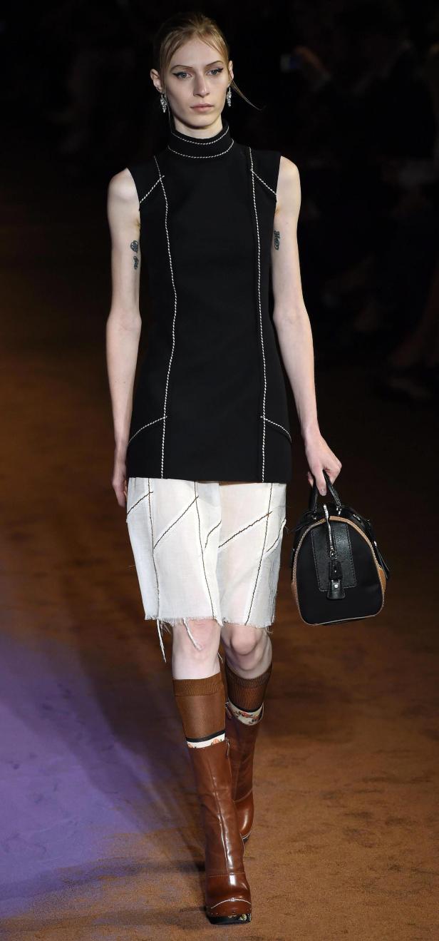 Eine Frau mit Tätowierungen trägt ein schwarzes Kleid, Shorts und braune Stiefel und hält eine Handtasche.