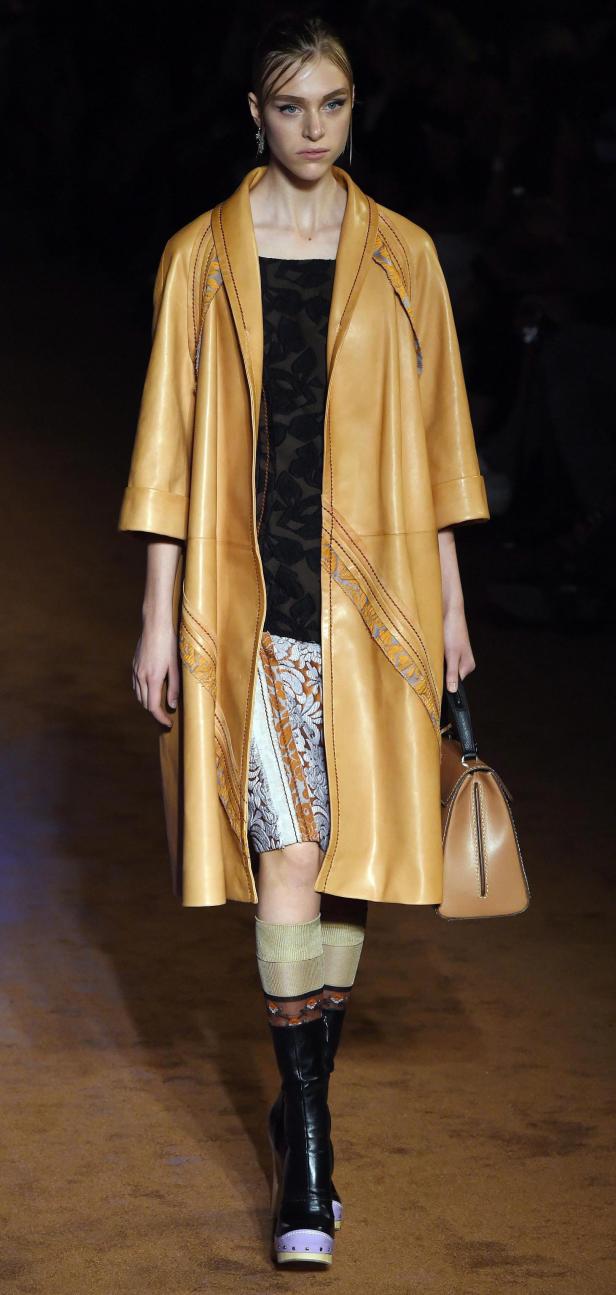 Eine Frau in einem hellbraunen Ledermantel mit passender Handtasche auf einem Laufsteg.