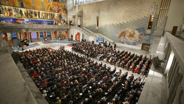 Eine große Menschenmenge sitzt in einem Saal mit Wandmalereien und einer Bühne.
