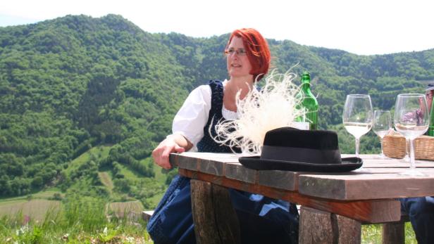 Eine Frau in Tracht sitzt an einem Tisch mit Wein und einem Hut vor einer grünen Hügellandschaft.