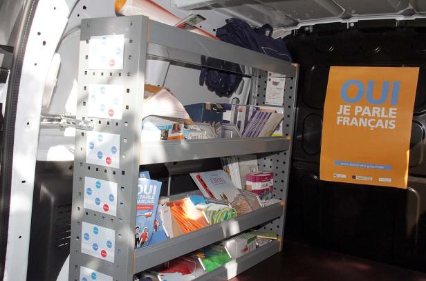 Ein Regal im Inneren eines Lieferwagens, gefüllt mit Broschüren und Flyern, darunter ein Poster mit der Aufschrift „Oui, je parle français“.