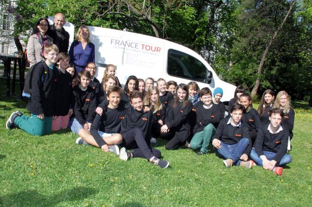 Eine Gruppe von Jugendlichen posiert vor einem „France Tour“-Bus auf einer Wiese.