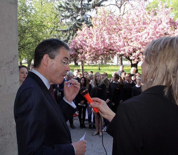 Ein Mann in Anzug spricht in ein Mikrofon vor einer Gruppe von Jugendlichen im Freien.