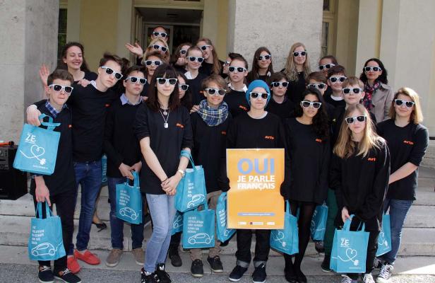 Eine Gruppe Jugendlicher mit Sonnenbrillen posiert mit blauen Taschen und einem Schild mit der Aufschrift „Oui je parle français“.