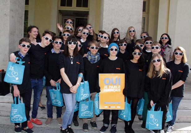 Eine Gruppe von Jugendlichen mit Sonnenbrillen posiert mit blauen Taschen und einem Schild „Oui je parle français“.