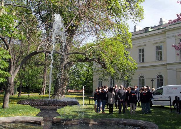 Eine Gruppe von Menschen steht vor einem Brunnen in einem Park, im Hintergrund ein helles Gebäude.