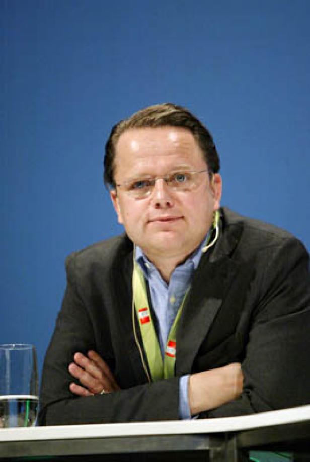 Ein Mann mit Brille und Anzug sitzt mit verschränkten Armen vor einem blauen Hintergrund.