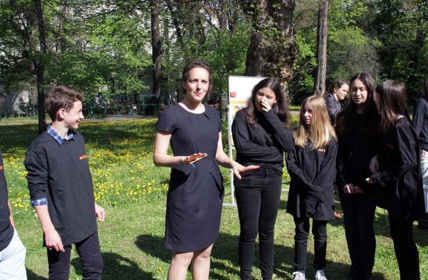 Eine Frau in einem schwarzen Kleid spricht vor einer Gruppe von Jugendlichen im Freien.