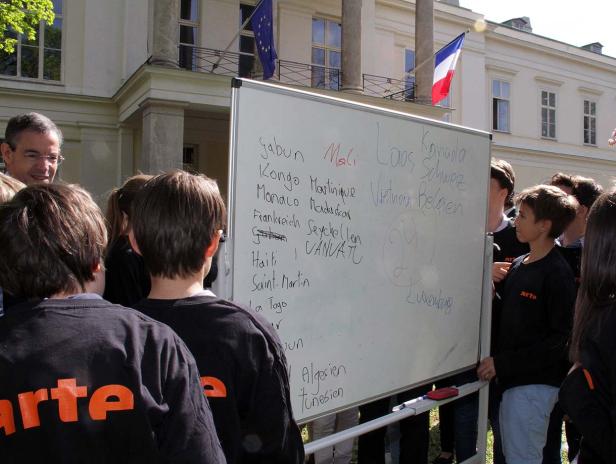 Eine Gruppe von Kindern steht vor einer Tafel mit Ländernamen vor einem Gebäude mit EU- und Frankreich-Flagge.