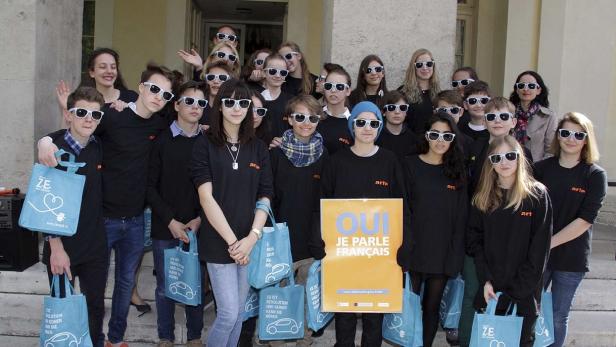 Eine Gruppe Jugendlicher mit Sonnenbrillen hält blaue Taschen und ein Schild mit der Aufschrift „Oui, je parle français“.