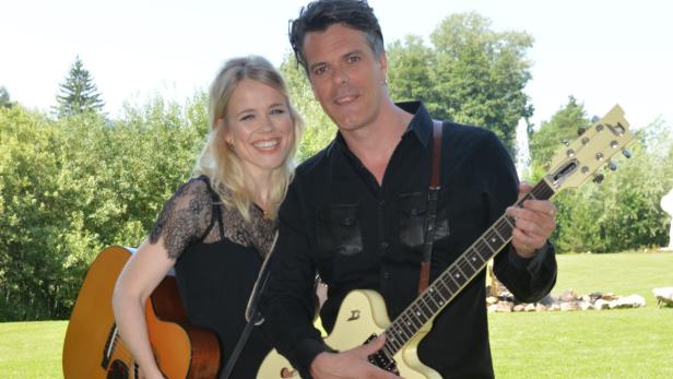 Eine Frau mit Gitarre und ein Mann mit E-Gitarre stehen lächelnd im Freien.