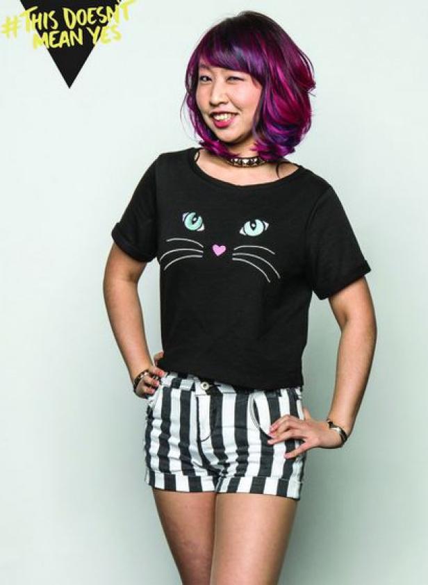 Eine Frau mit lila Haaren und einem schwarzen T-Shirt mit Katzenmotiv blinzelt in die Kamera.