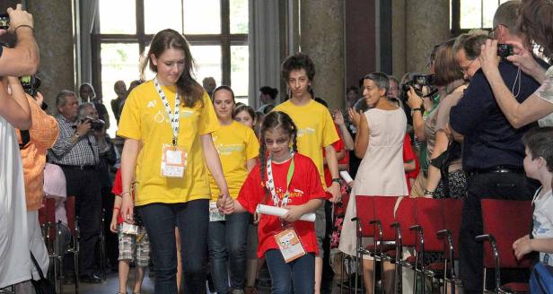 Eine Gruppe Kinder geht durch einen Raum, umringt von applaudierenden und fotografierenden Menschen.