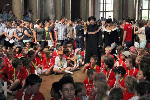 Eine Gruppe von Kindern sitzt vor zwei Personen in traditionellen akademischen Gewändern.
