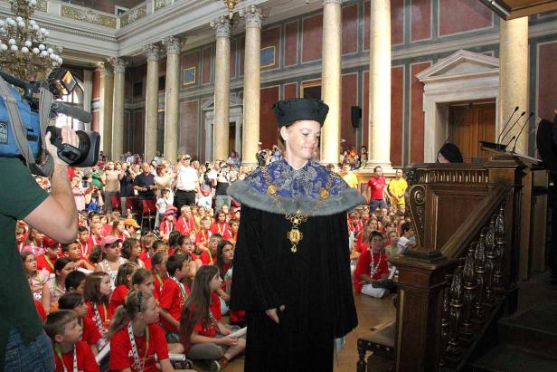 Eine Frau in akademischer Kleidung steht vor einer Gruppe von Kindern in einem großen Saal.