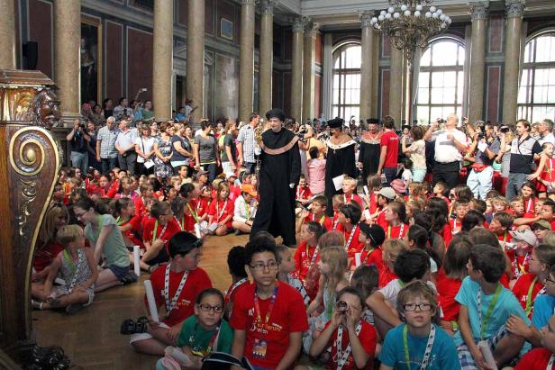 Eine Gruppe von Kindern sitzt auf dem Boden in einem großen Saal, während Erwachsene zusehen.