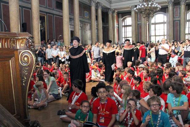 Eine Gruppe von Kindern sitzt vor drei Personen in akademischer Kleidung in einem großen Saal.