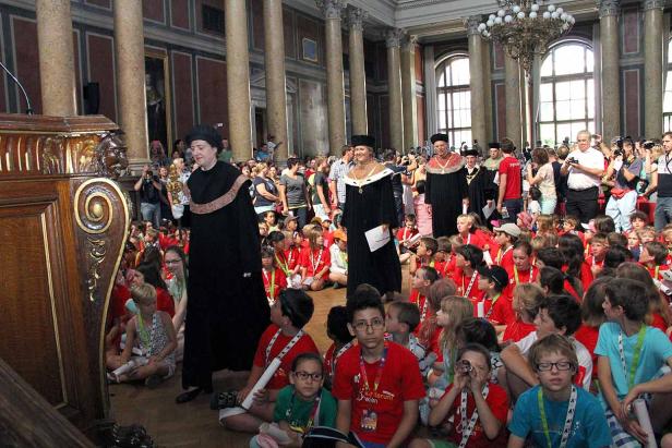 Eine Gruppe von Kindern sitzt vor einer Gruppe von Personen in akademischer Kleidung in einem großen Saal.