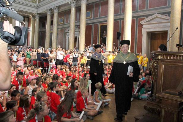 Eine Gruppe Kinder sitzt auf dem Boden, während zwei Personen in akademischer Kleidung vorbeigehen.