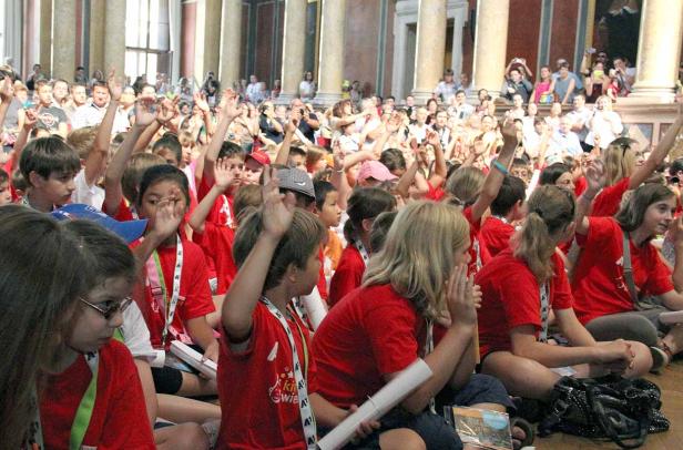 Eine große Gruppe Kinder in roten T-Shirts sitzt und hebt die Hände.