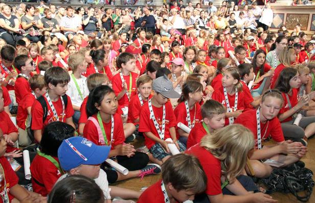 Eine große Gruppe Kinder sitzt auf dem Boden und schaut auf eine Bühne mit Publikum im Hintergrund.