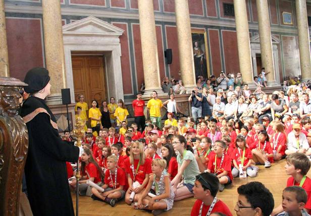 Eine Gruppe Kinder sitzt auf dem Boden in einem großen Saal und hört aufmerksam zu.