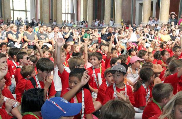 Eine große Gruppe Kinder in roten T-Shirts sitzt in einem Saal und meldet sich.