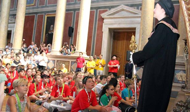 Eine Gruppe Kinder sitzt auf dem Boden und hört einer Frau in historischer Kleidung zu.