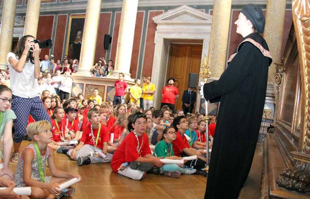 Eine Gruppe Kinder sitzt auf dem Boden und hört einer Person in einer Robe zu.