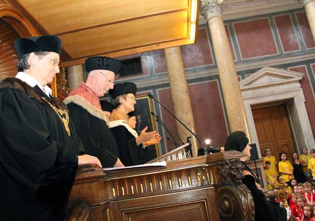 Mehrere Personen in akademischer Kleidung stehen an einem Rednerpult in einem Saal.