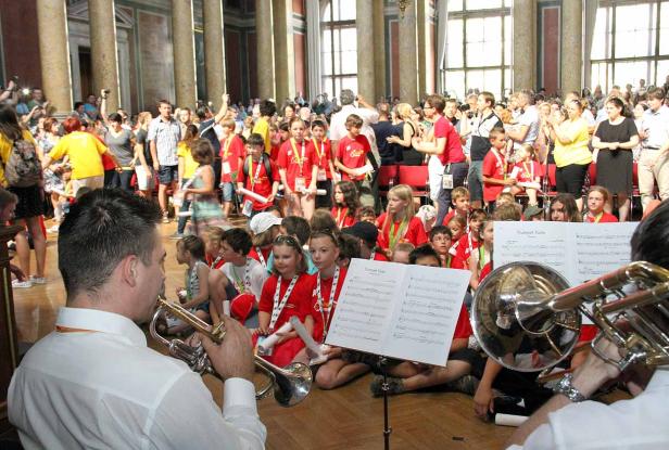 Eine Gruppe Kinder in roten T-Shirts sitzt auf dem Boden und spielt Trompete vor einem Publikum.