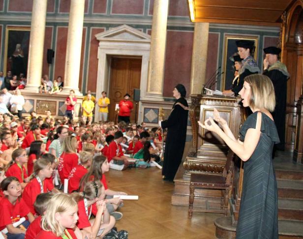 Eine Frau hält eine Rede vor einer Gruppe von Kindern in einem großen Saal.