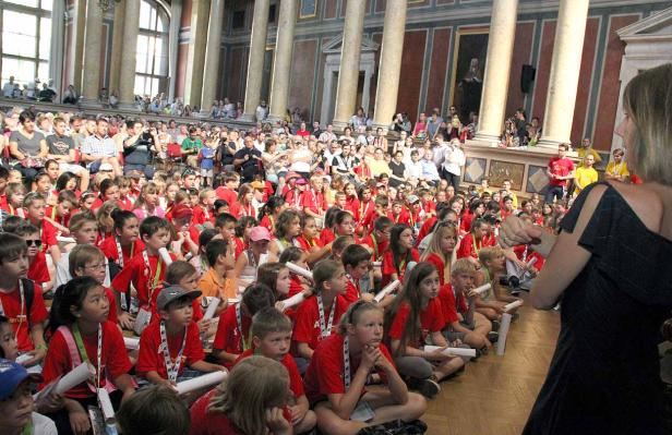 Eine große Gruppe Kinder sitzt auf dem Boden in einem Saal und hört aufmerksam zu.